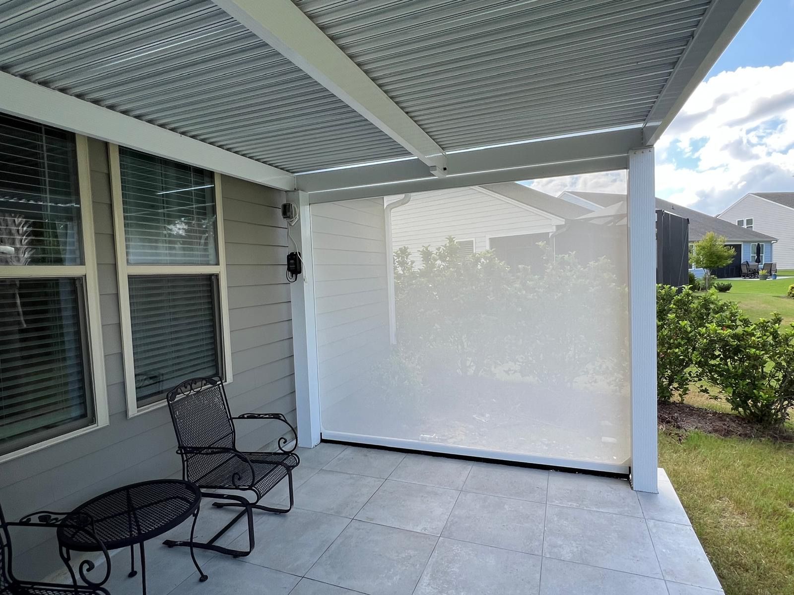A patio with a light gray tiled floor, a metal chair, a side table, a corrugated metal roof, and a beige privacy screen.