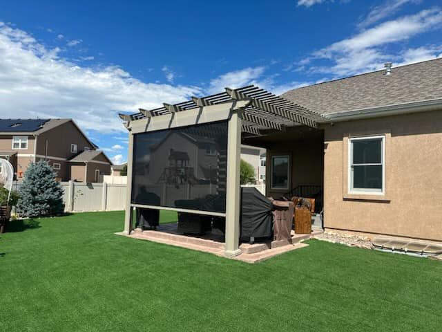 A tan pergola with a lowered dark privacy screen over a brick patio in a residential backyard with green grass.