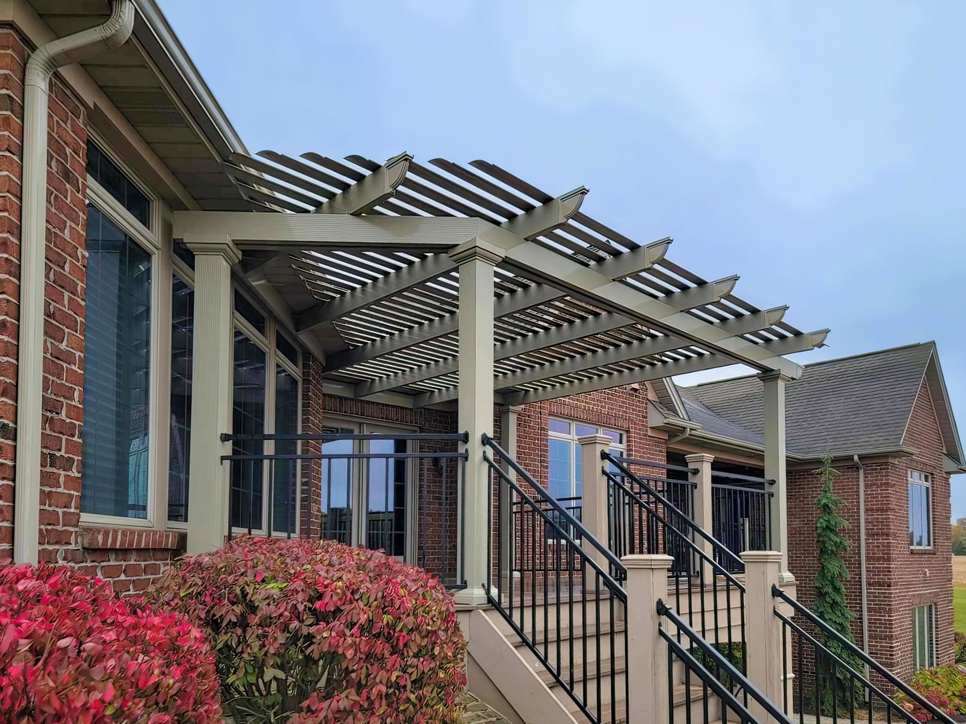 A brick house exterior with a wooden pergola covering a patio staircase featuring black metal railings and landscaping.