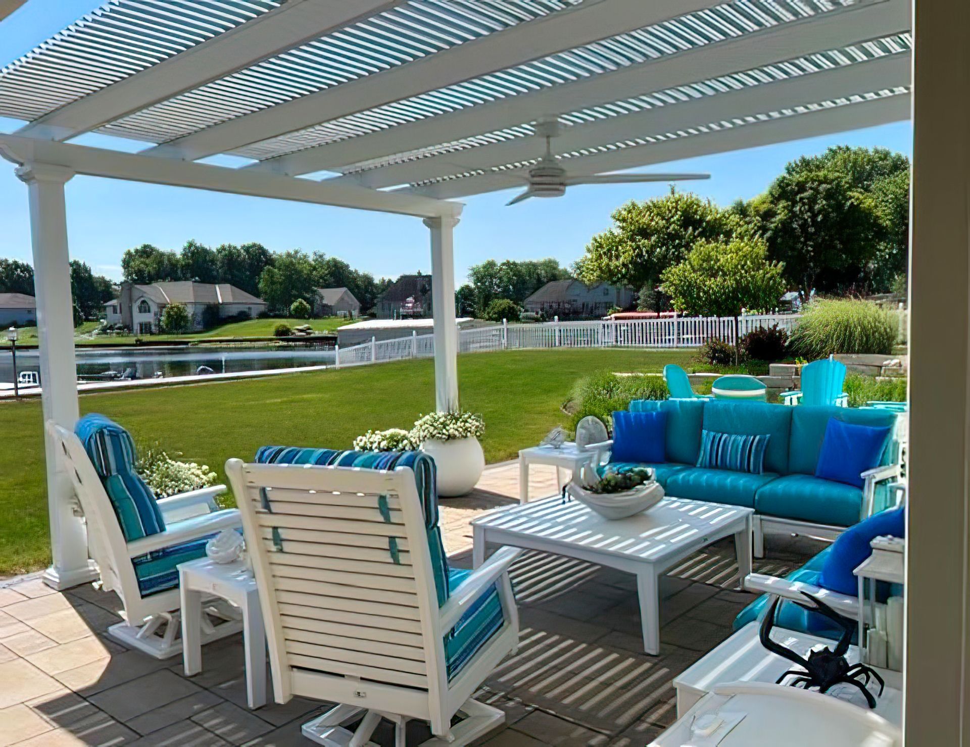 A patio with a white pergola and ceiling fan, featuring a blue sofa and chairs overlooking a grassy lawn and a pond.