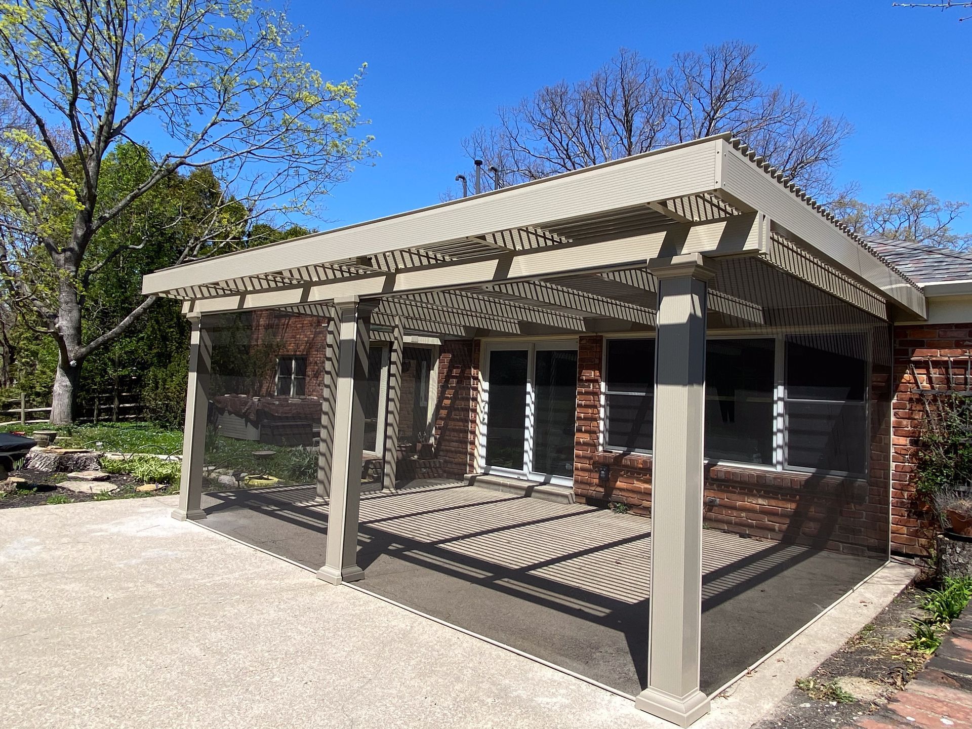 A beige pergola with a lattice roof attached to a red brick house, shading a concrete patio on a sunny day.