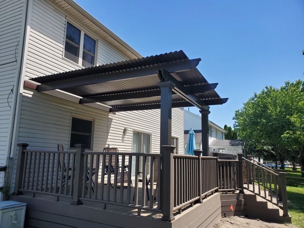 A brown wooden deck with a matching pergola and corrugated roof attached to the side of a light-colored house.