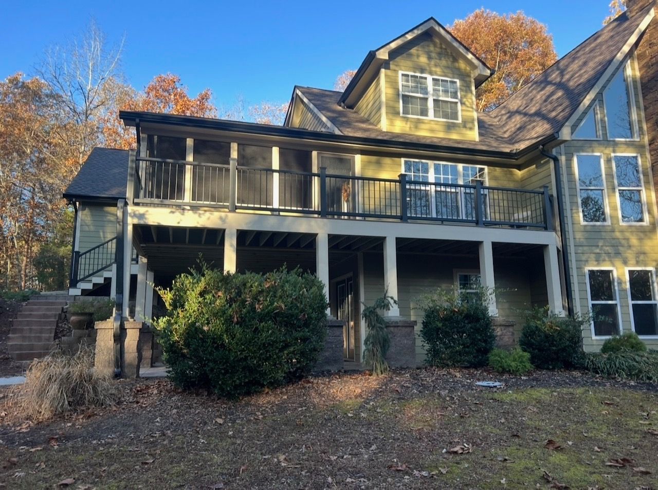 Two-story house with green siding and black railings. Trees in the background, deck, and shrubbery in front.