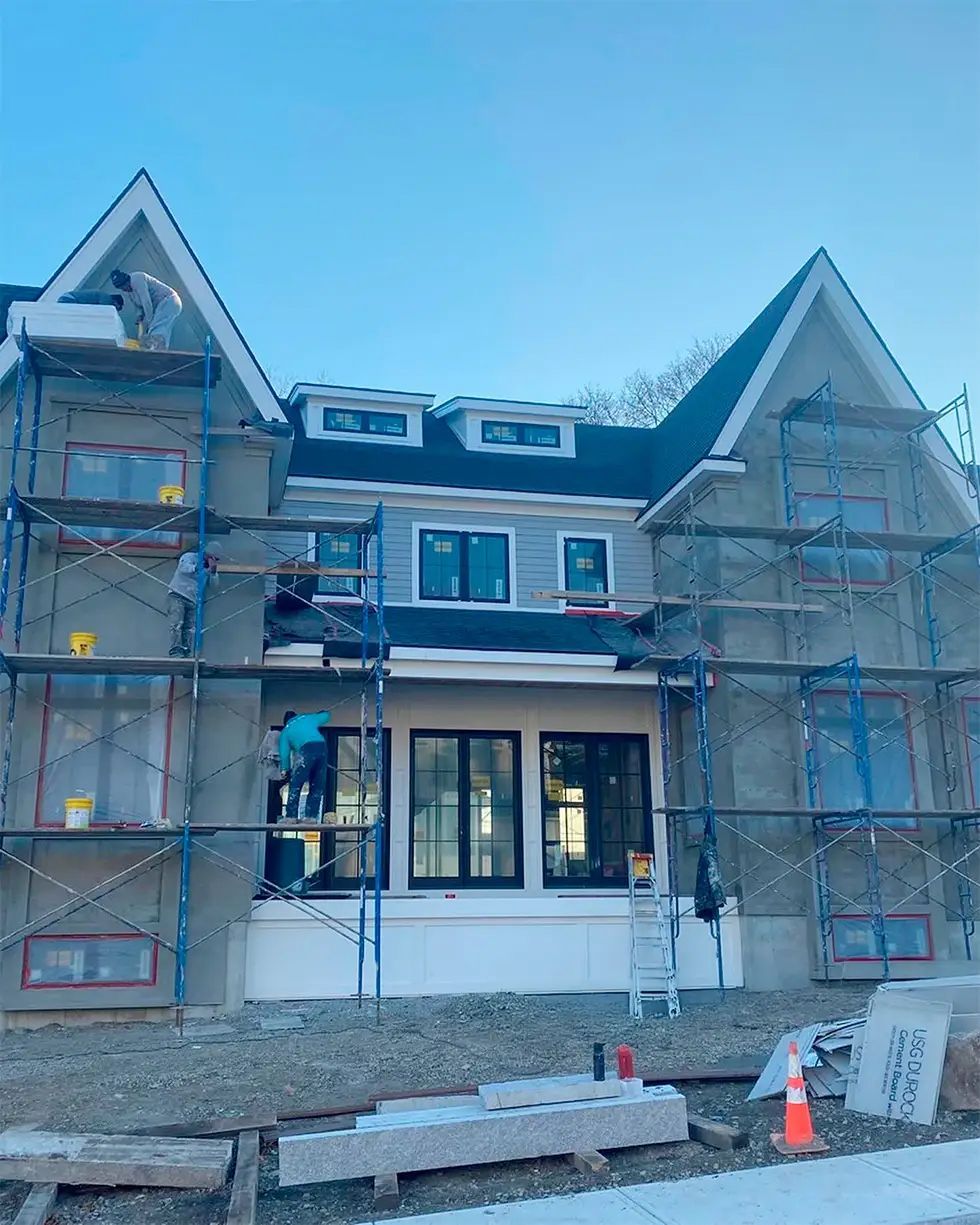 Construction workers on scaffolding, plastering the exterior of a two-story house with gray siding.