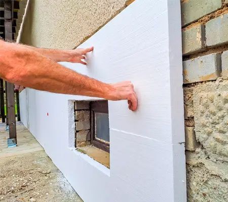 Person installing white insulation board on a brick wall with a small window.