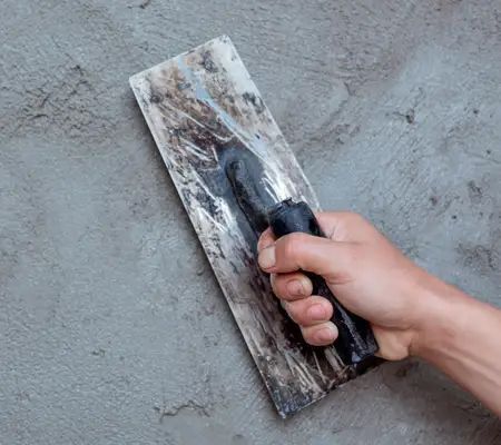 Hand holding a trowel smoothing grey plaster on a wall.