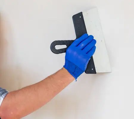 Person wearing blue gloves, using a putty knife on a white wall.