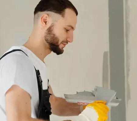 Man in overalls smoothing wall with a trowel, wearing gloves.