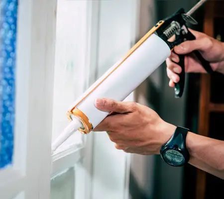 Person caulking a white window frame with a caulk gun.