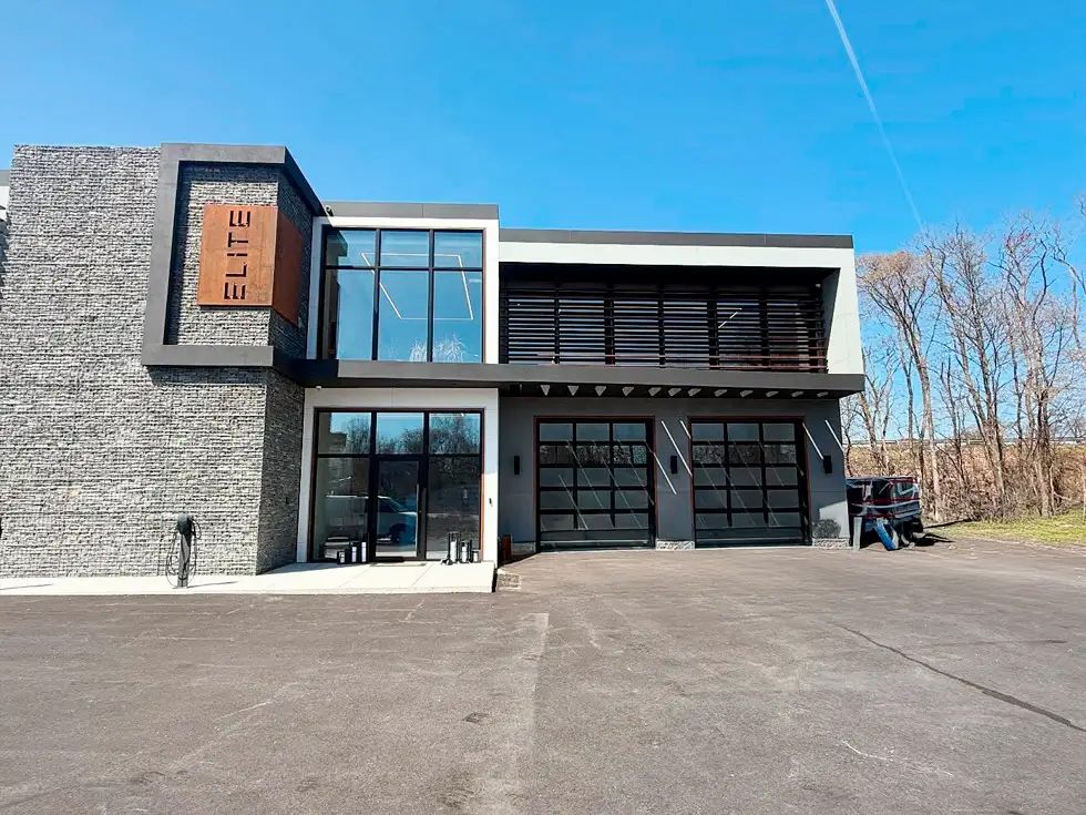 Modern building with textured gray facade, glass windows, and two garage doors under a clear blue sky.