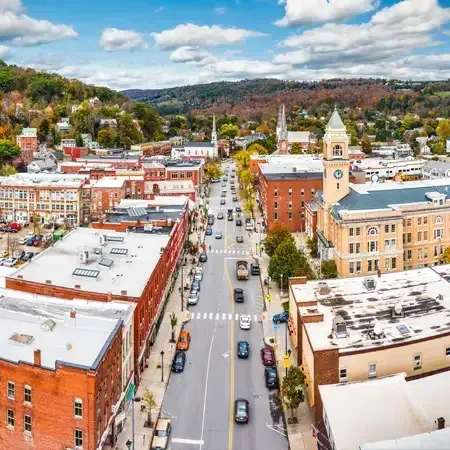 Aerial view of a town with a long street lined with buildings and cars; autumn trees in the background.