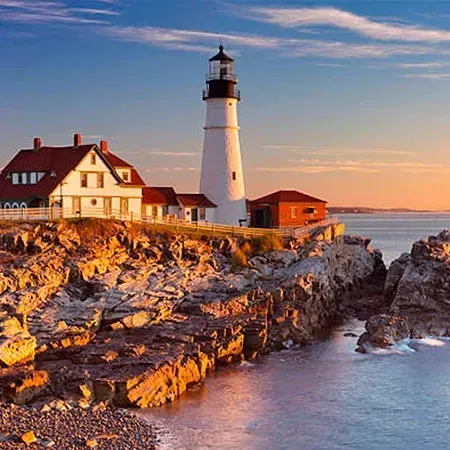 Lighthouse on rocky coast at sunset; white tower, red roof buildings, ocean, blue sky.