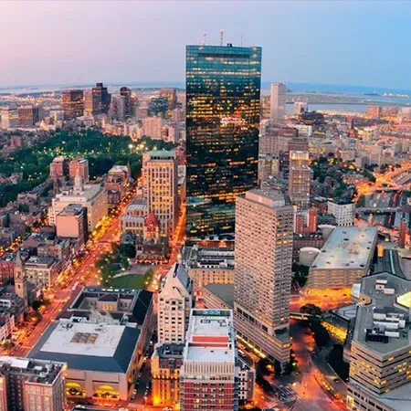 City skyline at dusk, Boston, MA. Tall buildings with lit windows, roadways, and a harbor in the distance.
