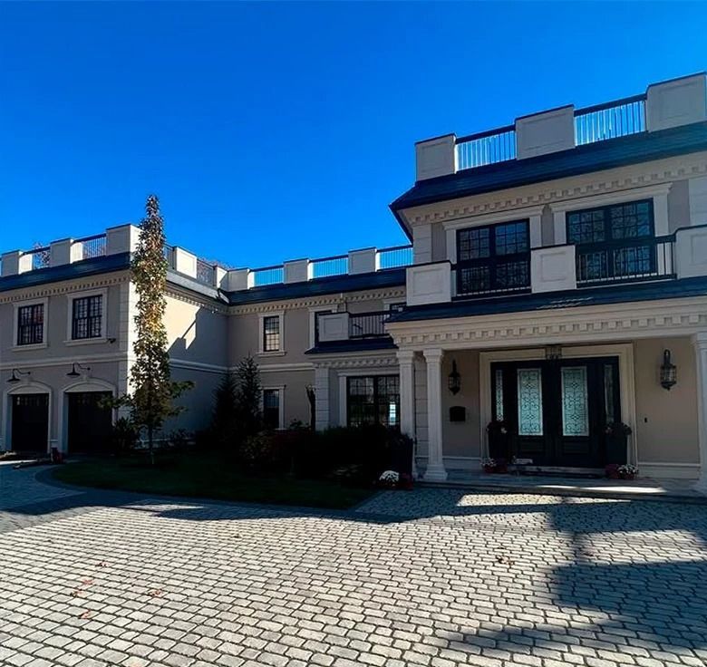 Large light-colored house with black doors, balcony, and stone driveway under a blue sky.