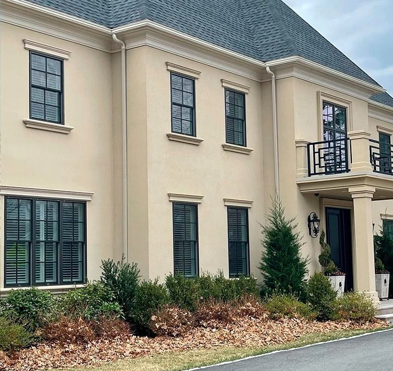 Beige stucco house with dark windows and shutters, black balcony, and green shrubs.