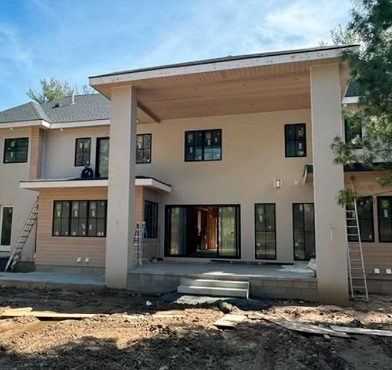 Rear exterior of a house under construction with a covered patio. Beige stucco walls, black windows, and a partially finished yard.