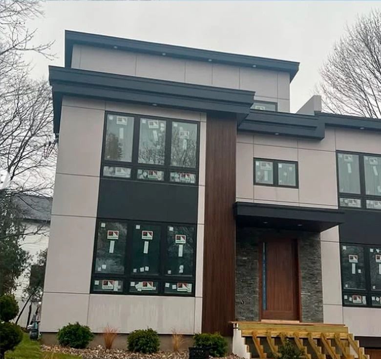 Modern two-story house with gray and brown exterior. Large windows, wooden door, and stone accents.