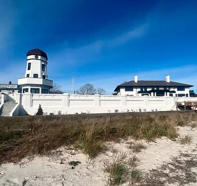 White building with a tower, a low wall, and a dark roof, on a sandy beach under a blue sky.