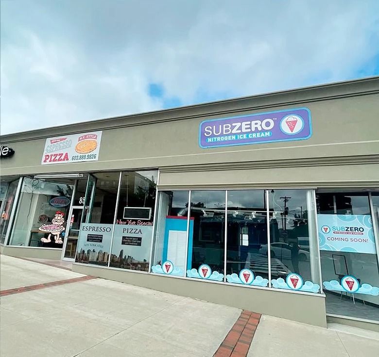 Exterior of a strip mall with a pizza shop and Sub Zero ice cream shop under a cloudy sky.