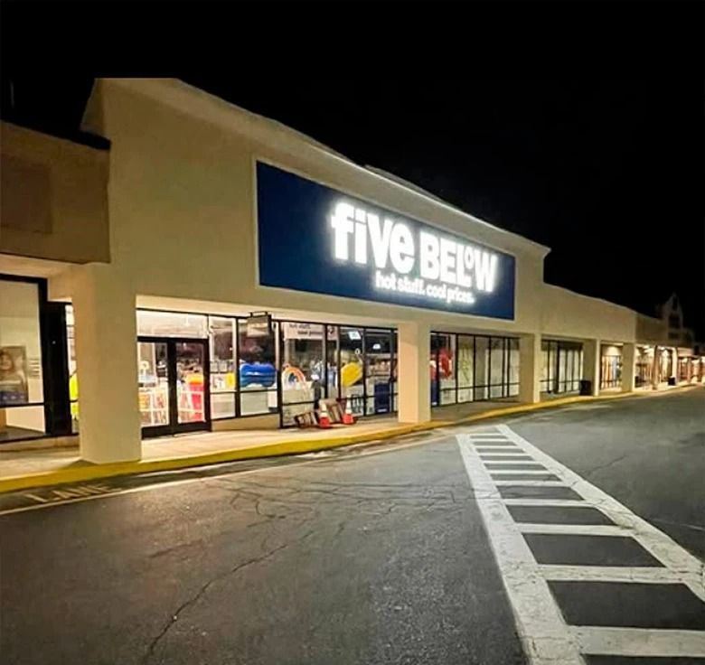 Exterior of a Five Below store at night, with illuminated sign and merchandise visible through windows.
