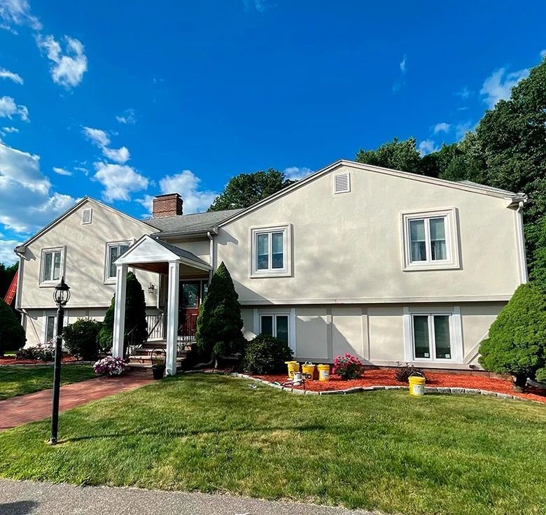 Two-story house with light beige facade, red brick walkway, green lawn, and blue sky.