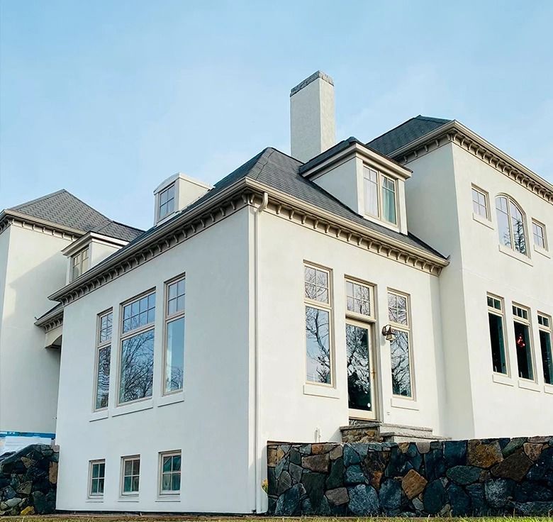 White multi-story house with dark roof and chimney, stone wall in front.