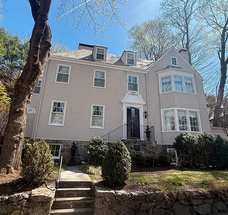 Gray stucco house with white trim, bay windows, and a stone retaining wall.