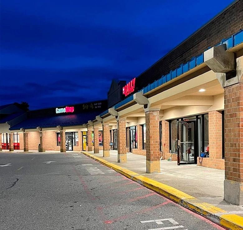Strip mall at dusk with stores like GameStop and Sally's. Dark blue sky, red brick, and storefronts.