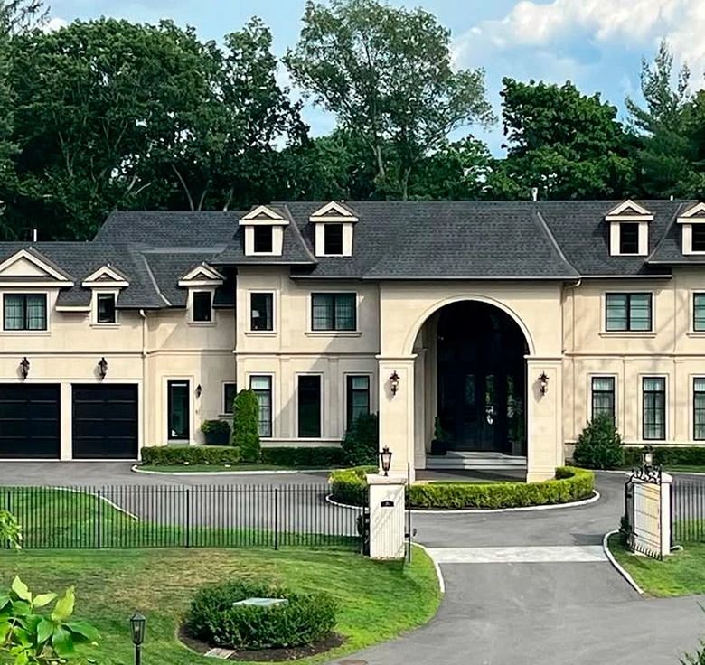 Luxurious beige home with black roof, multiple dormers, and arched entryway. Black garage doors and manicured lawn.
