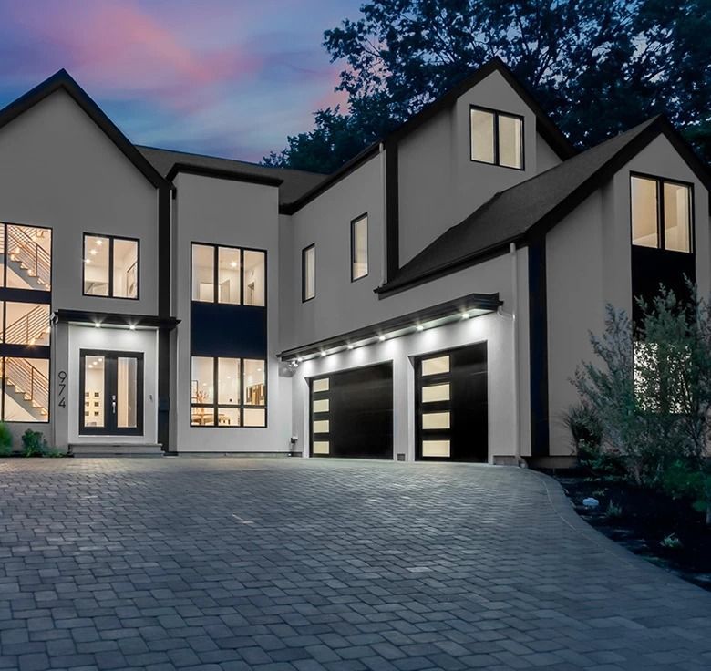 Modern white house with black trim, two-car garage, and a cobblestone driveway at dusk.