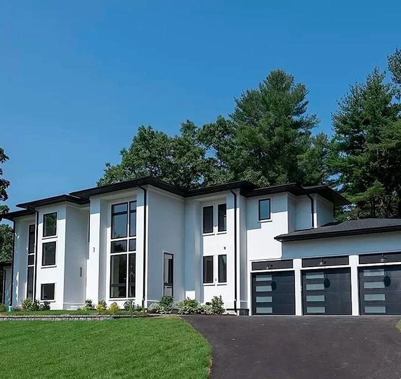 Modern white house with black trim, black garage doors, and a curved driveway on a green lawn. Blue sky.