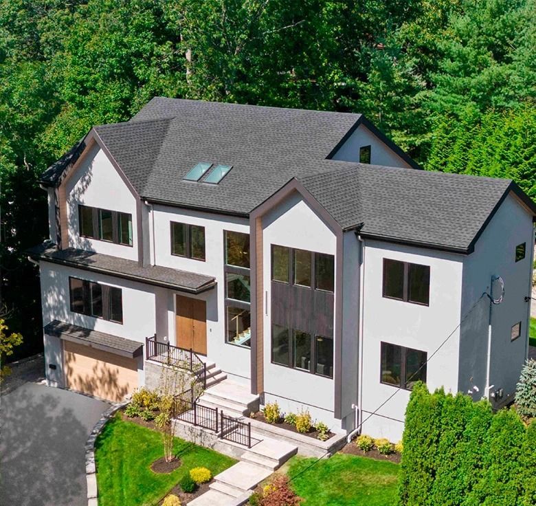 Modern two-story house with gray stucco walls, dark roof, multiple windows, and a landscaped front yard.