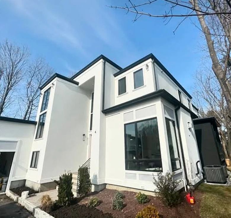 Modern, white two-story house with black trim and windows, set against a blue sky, surrounded by trees.