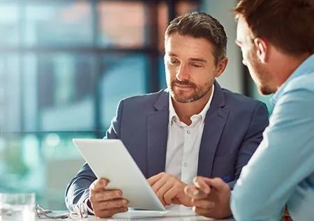 Two men in business attire review a tablet in an office setting.