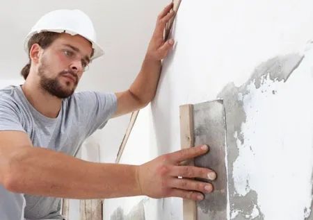 Man in hard hat applies plaster to a wall with a trowel.