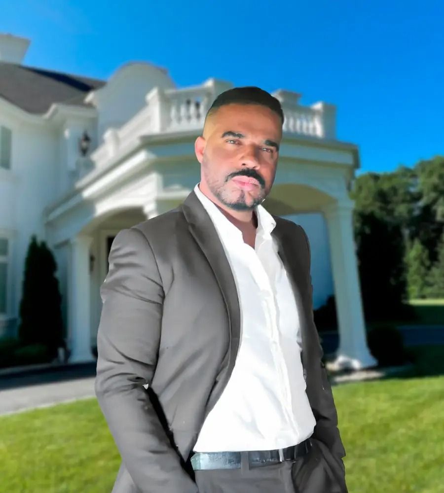 Man in a suit standing in front of a large white house with a green lawn.