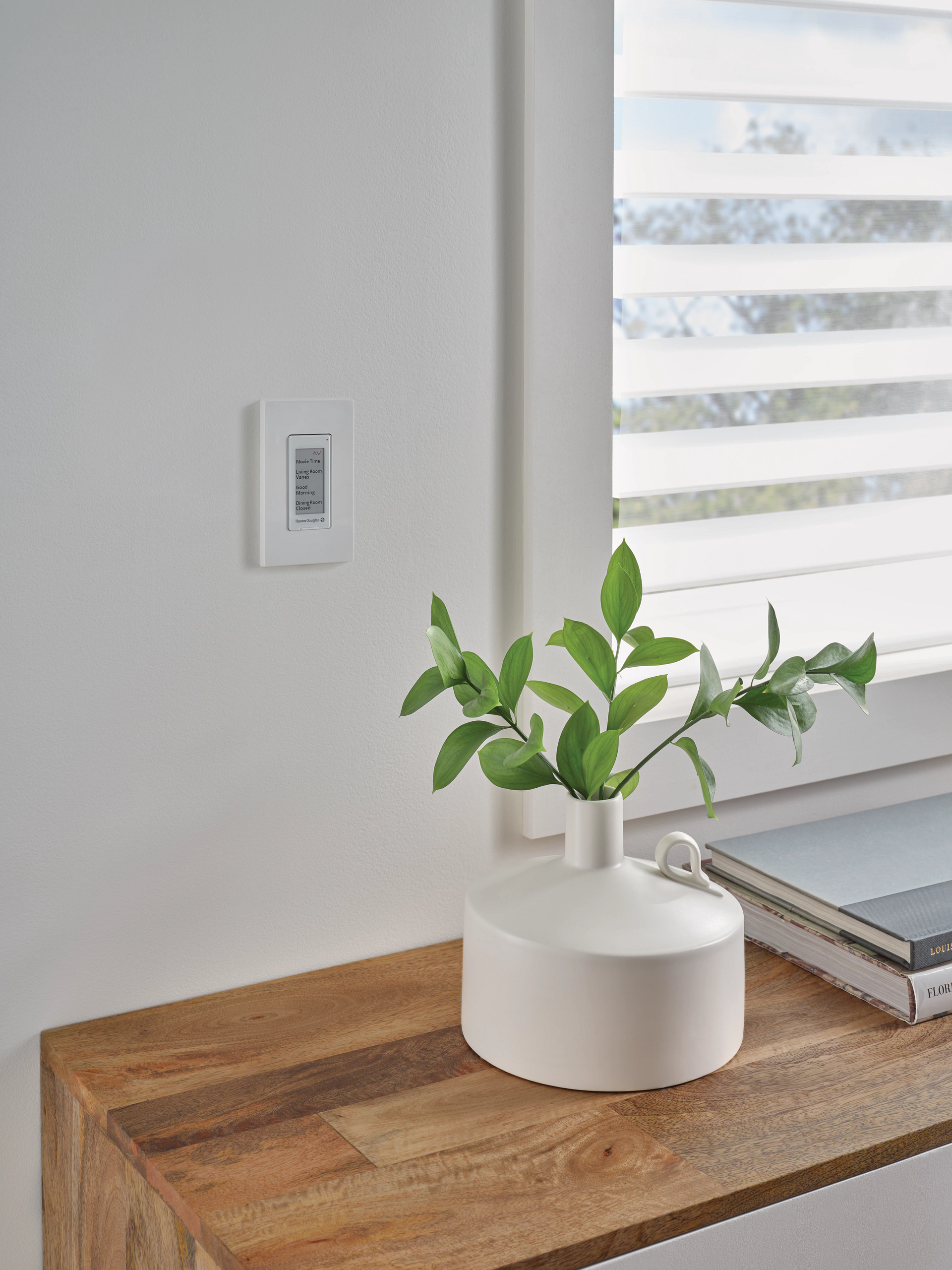 A white vase with a plant in it is sitting on a wooden table next to a window.