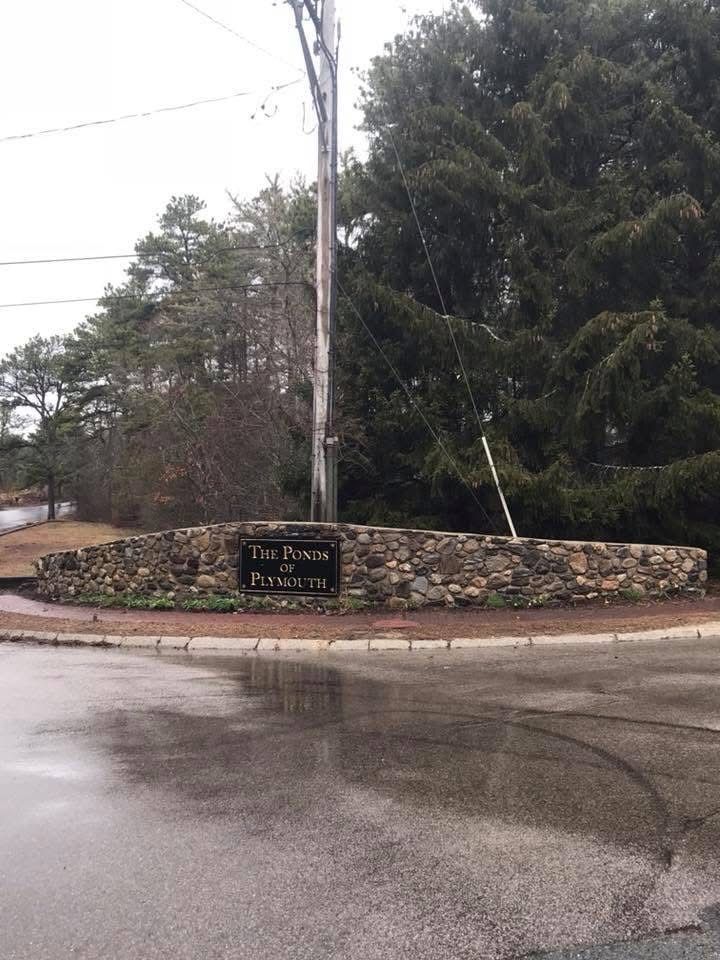 A stone sign reading The Ponds at Foxhollow at a street intersection, framed by pine trees under an overcast sky.