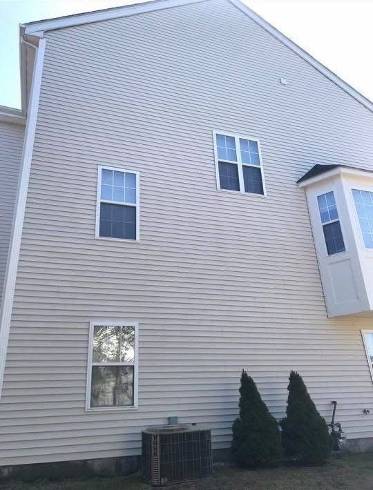 Side view of a beige-sided two-story house exterior with three windows, a bay window, and two small evergreen shrubs.