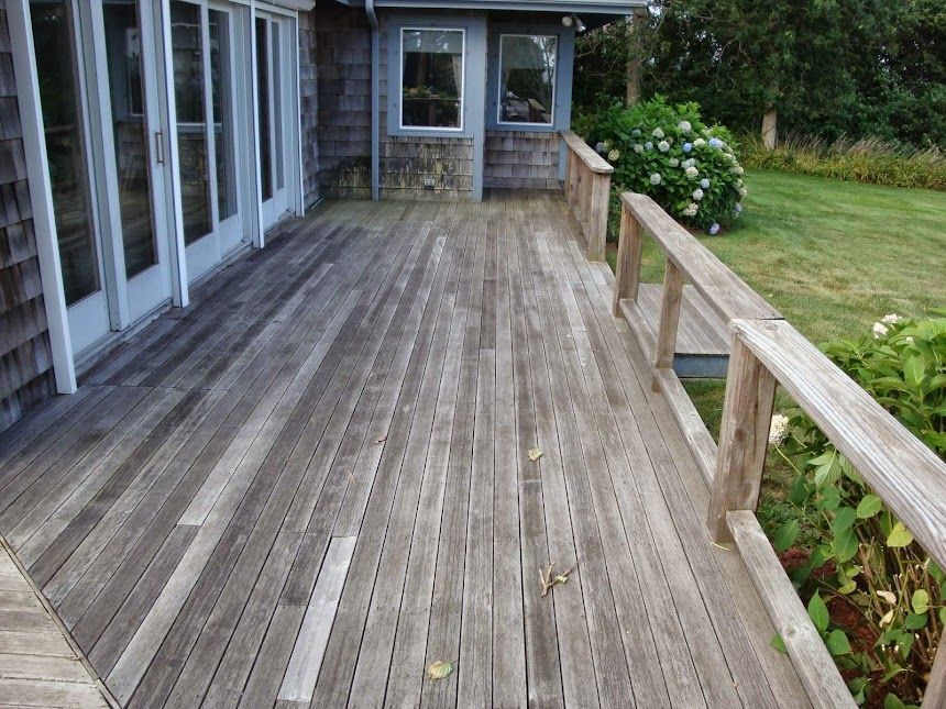 Weathered wooden deck attached to a house with sliding glass doors and a simple railing overlooking a green lawn.