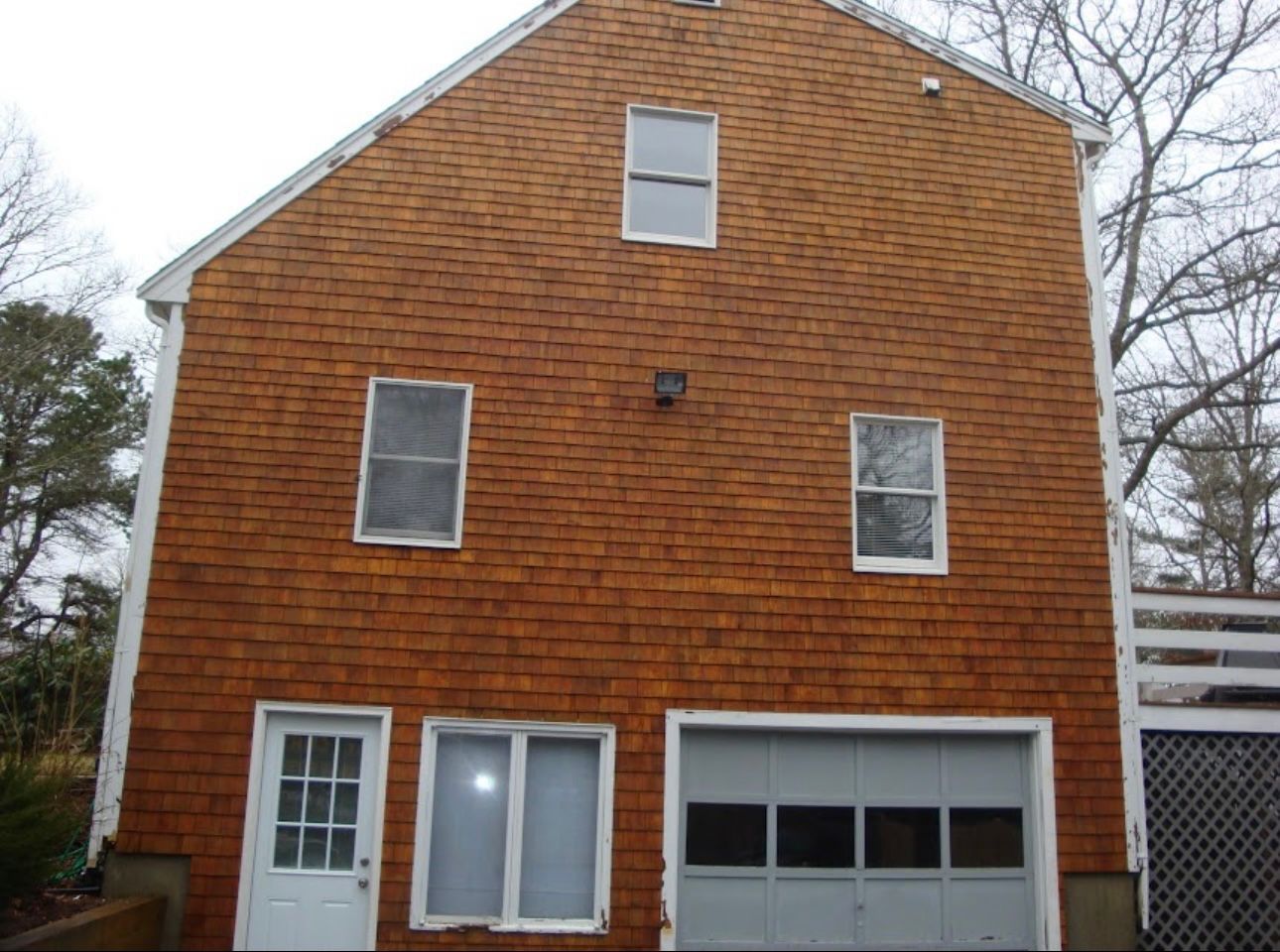 A brown shingled house exterior featuring a white door, windows, and a gray garage door under an overcast sky.