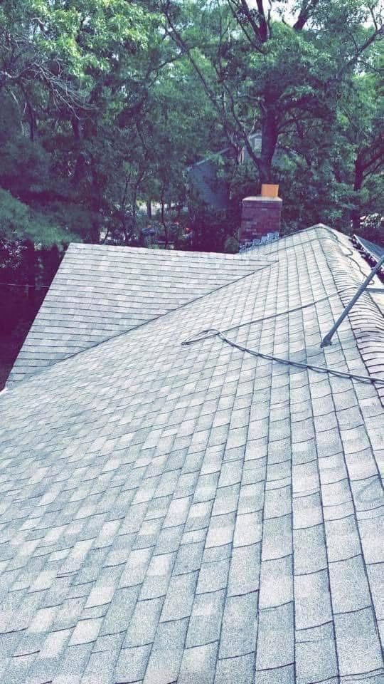An angled, high-view shot of a shingled roof with a brick chimney in a wooded setting.