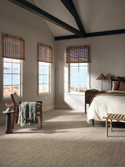 Bright bedroom with large windows, beige carpet, dark beams, and a bed with woven shades.