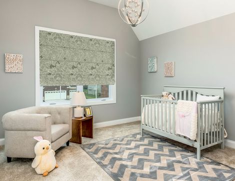 Nursery with gray walls, white crib, beige chair, chevron rug, and large window with patterned shade