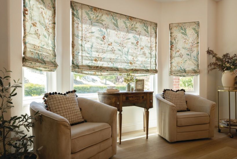Bright sitting room with two beige armchairs, floral blinds, and a small wooden table by large windows