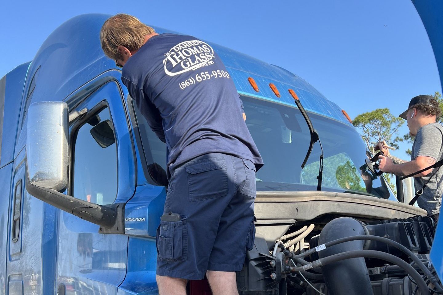 Two workers inspect a blue semi-truck’s front windshield under a clear sky.