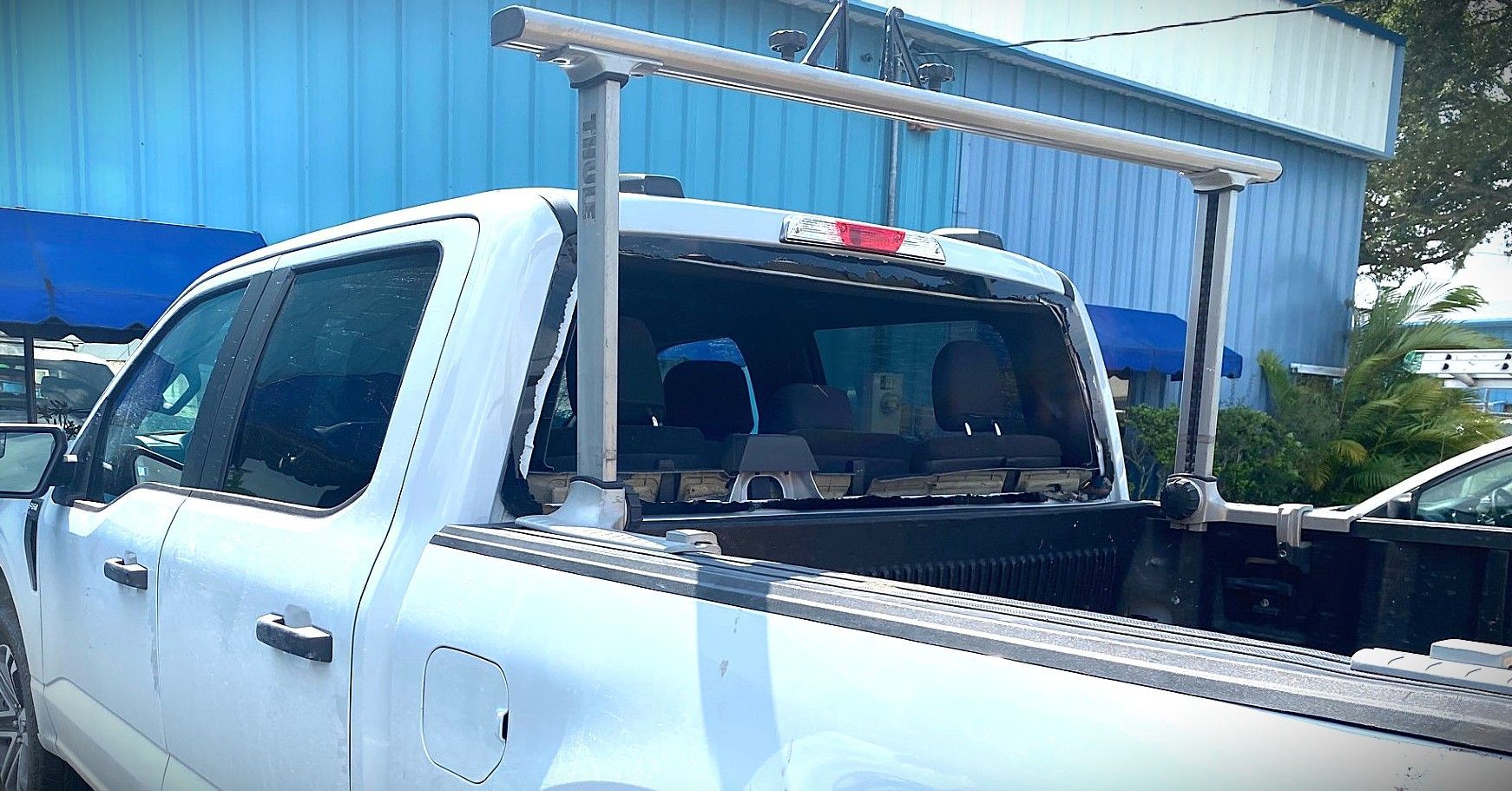 White pickup truck parked beside a blue building, carrying a ladder rack in the bed.