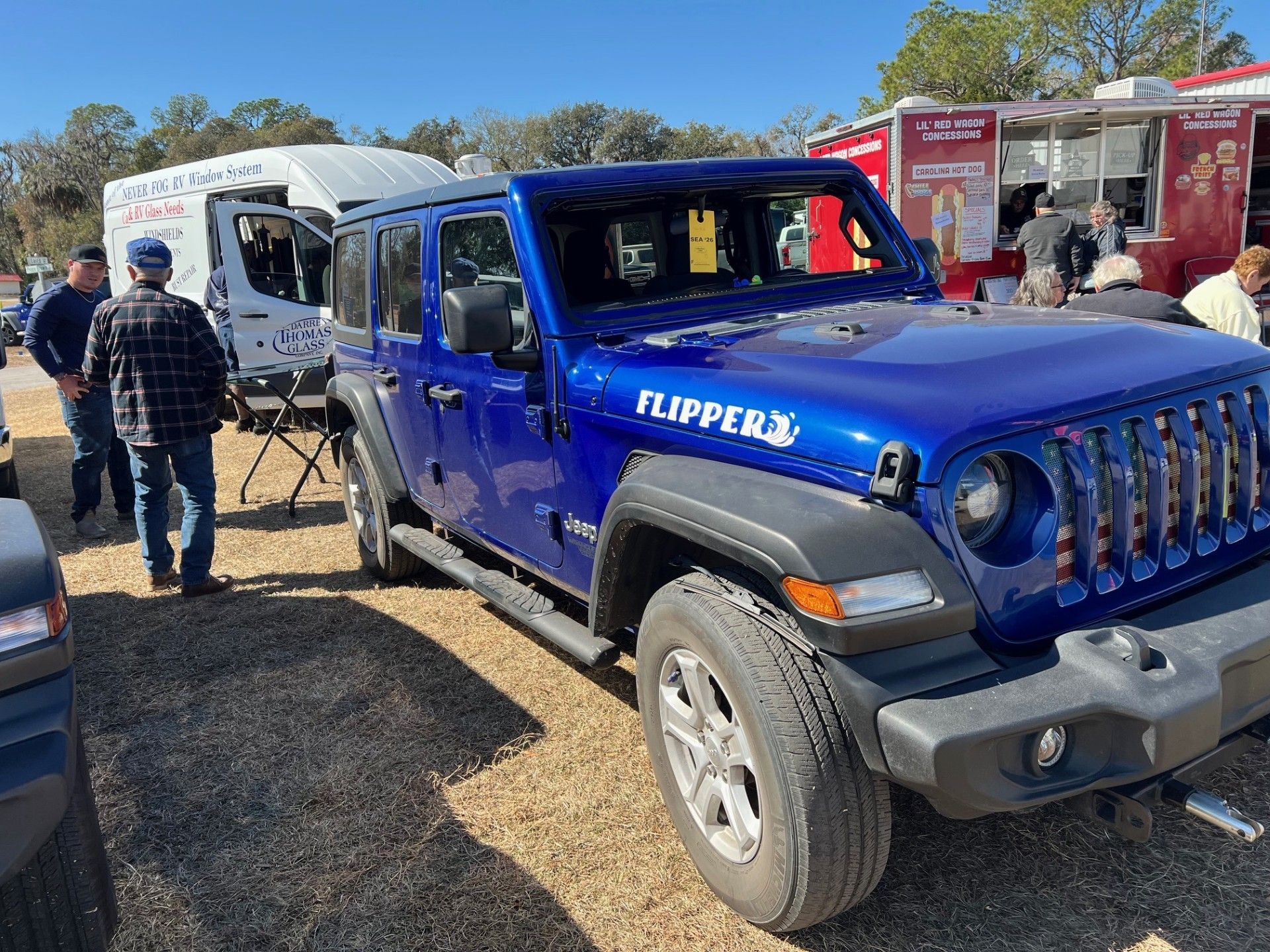 Blue Jeep Wrangler with “FLIPPER” lettering at an outdoor event, with people and trailers nearby