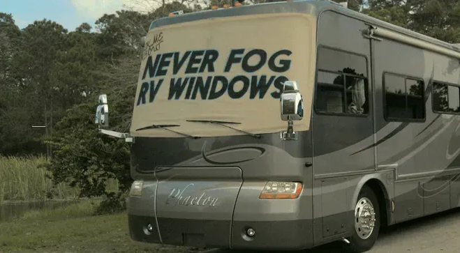 A large, gray recreational vehicle parked outdoors, featuring a banner on its windshield that reads "NEVER FOG RV WINDOWS."