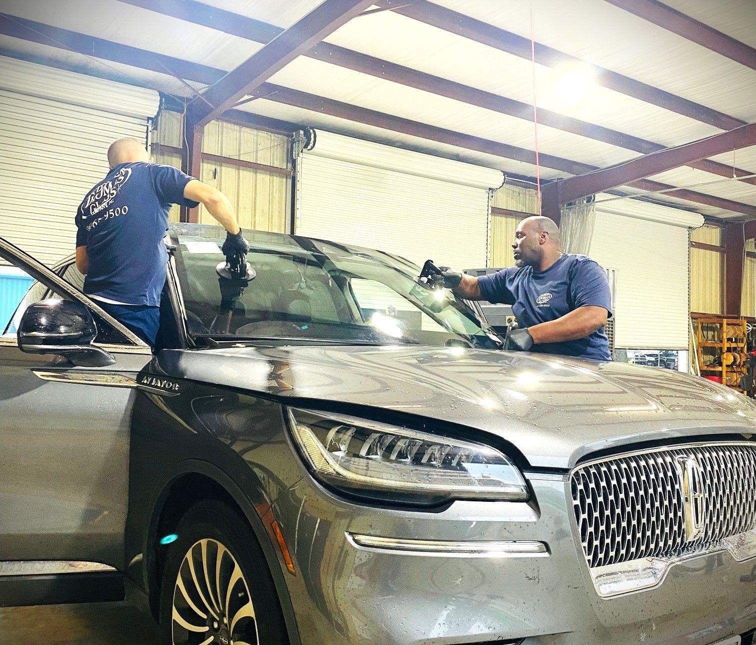 Two mechanics cleaning a silver SUV in a garage with open hood and windshield wipers raised